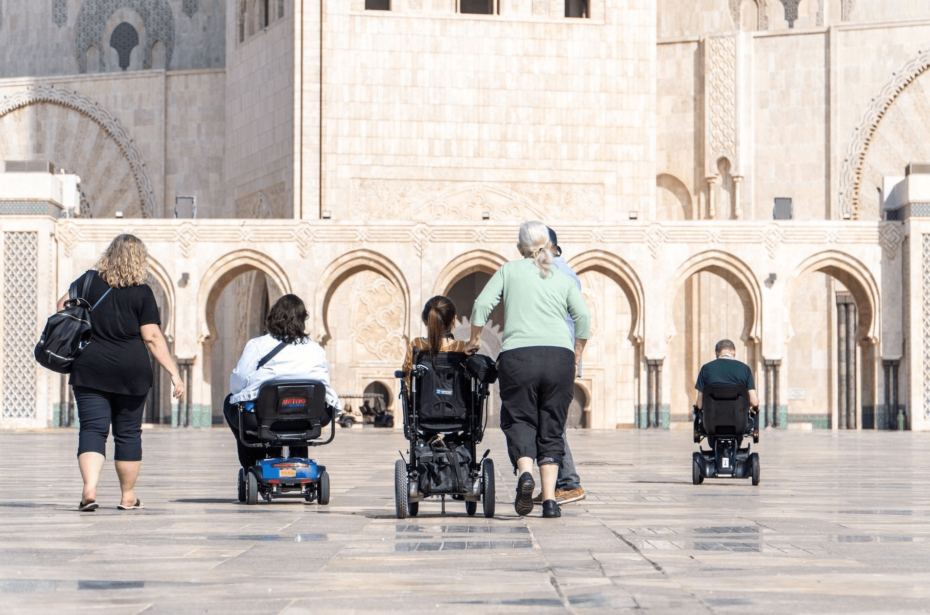 Three people in wheelchairs with three companions that are photographed with their backsides to the photo looking towards an old beige building with arched entrances