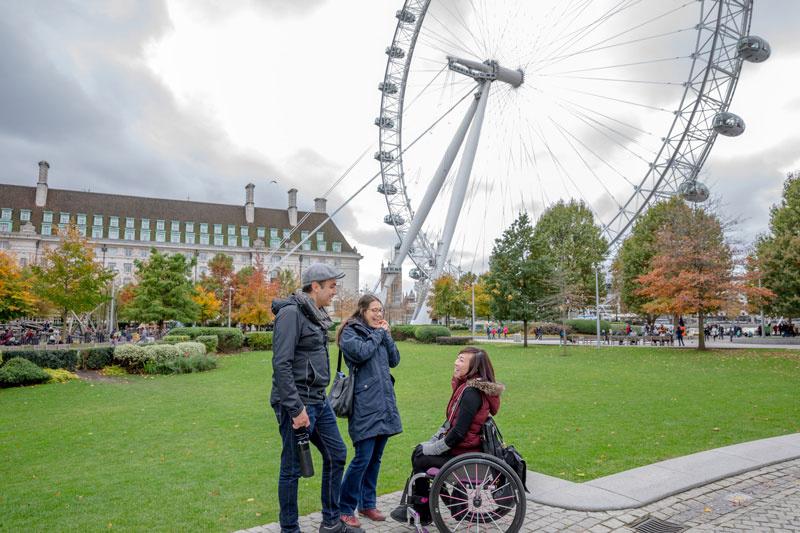 Three people, including a person in a wheelchair, behind the London Eye.