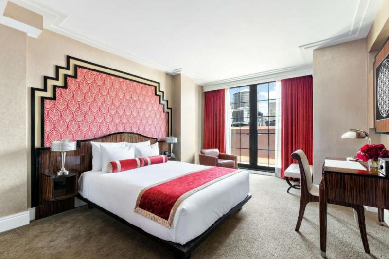Spacious room with wide space around the bed. Seating area by the window and access to a balcony. White and red linen and carpeted floor.