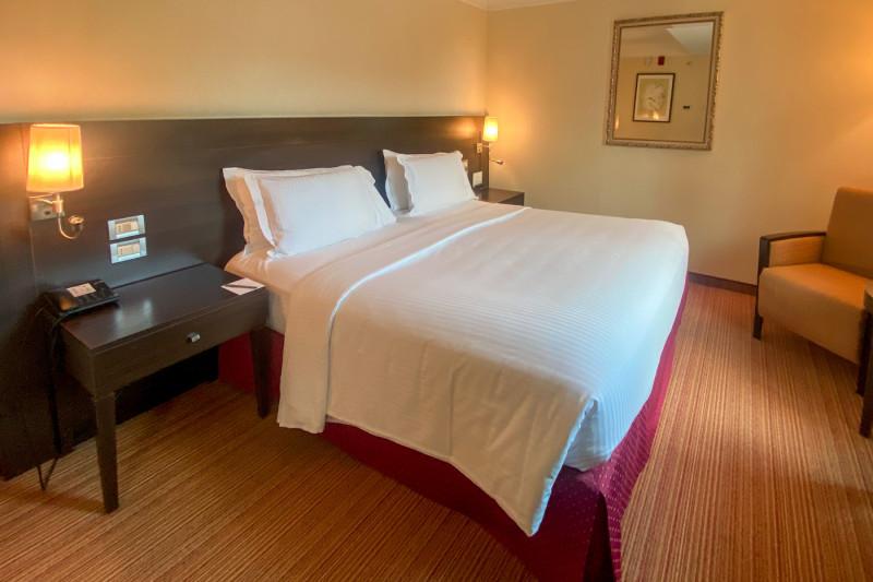 This smaller guest room has a queen-size bed, with nightstands on either side. Each table has a lamp mounted over it. There is a push button phone on one nightstand. The wall has a large mirror and there is a chair in the corner.