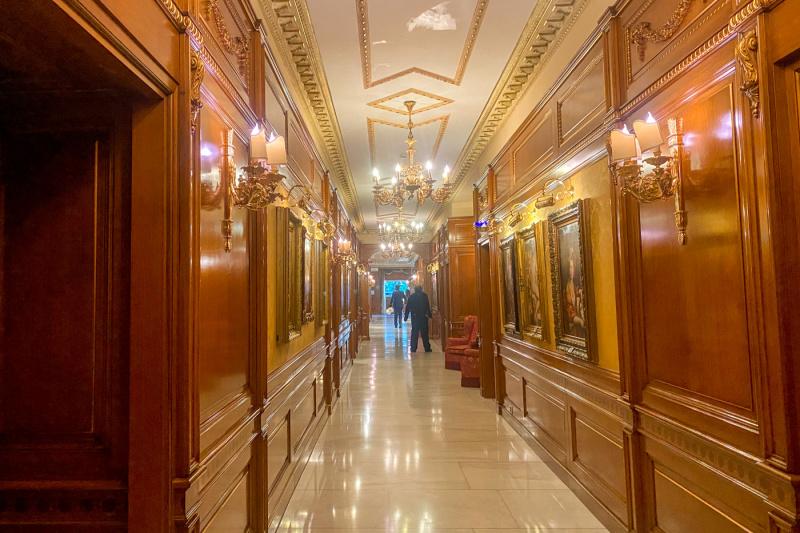 The wood-paneled corridor has chandelier lighting, artwork on the walls and a marble floor.