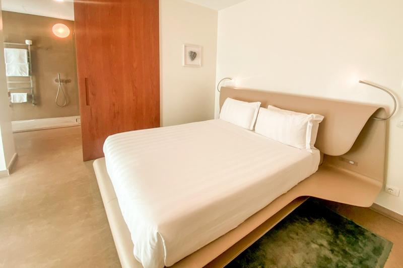 The room has a king-sized bed with white linen.The modern headboard has in-built nightstands and reading lamps. The ensuite is accessed through a sliding door.