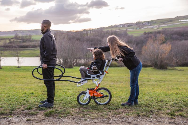 A joëlette kids wheelchair is a single wheeled wheelchair with support and steering bars in the front and back.