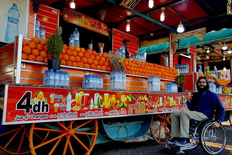 A man in a wheelchair smiles at the camera in front of a juice cart stocked with water bottles and oranges.