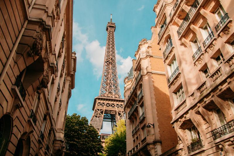 Looking up at the Eiffel Tower as it rises between two lines of typical Parisian buildings.