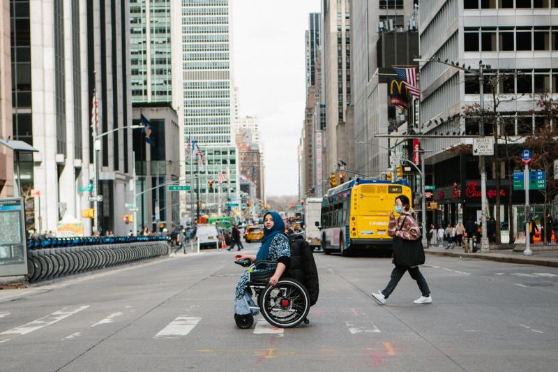 A person in an electric wheelchair crosses a busy New York street at a pedestrian crossing.
