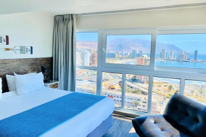 A guest room with a bed and a wall of windows that look out onto the city and ocean.