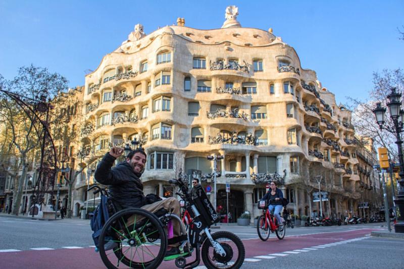 A man in a wheelchair smiles and waves in front of the La Pedrera building.