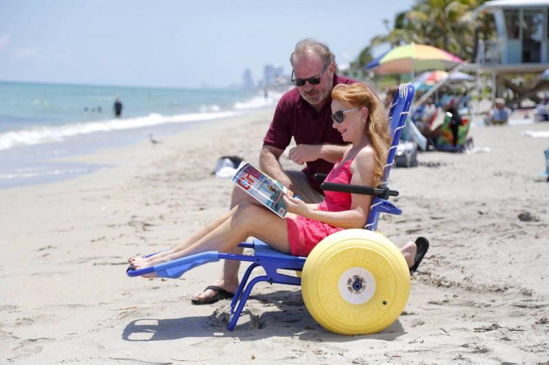 A person sits in a Joy on the Beach chair in an upright position, reading a magazine next to the sea.