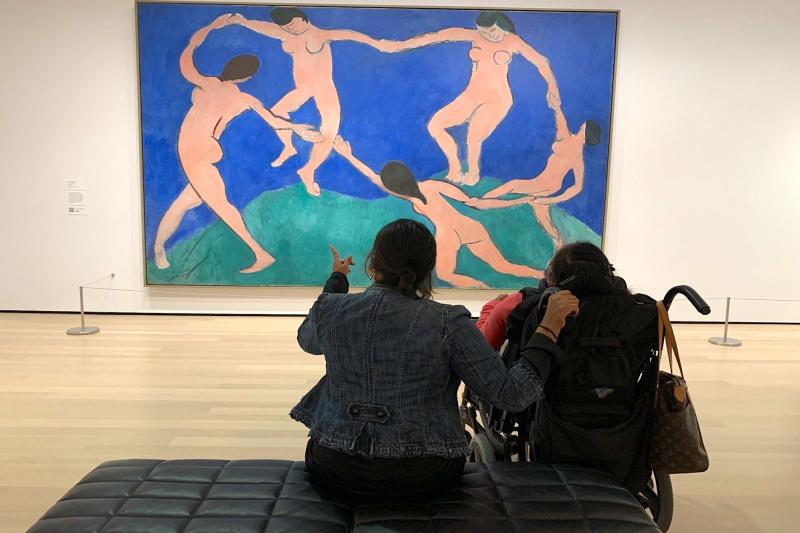 Two visitors, including a person in a wheelchair, look at a painting in the gallery.