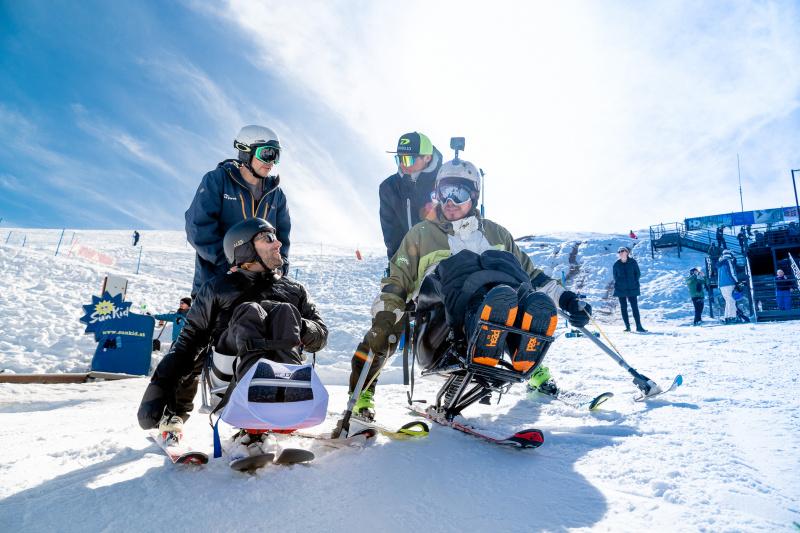 Two people in adaptive ski equipment on the slopes.